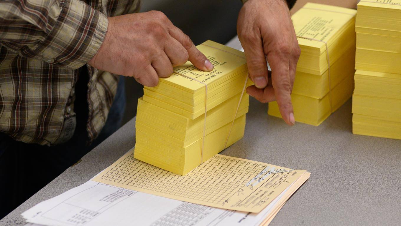 A person wearing a plaid shirt organizes several neat stacks of yellow paper sheets on a table.
