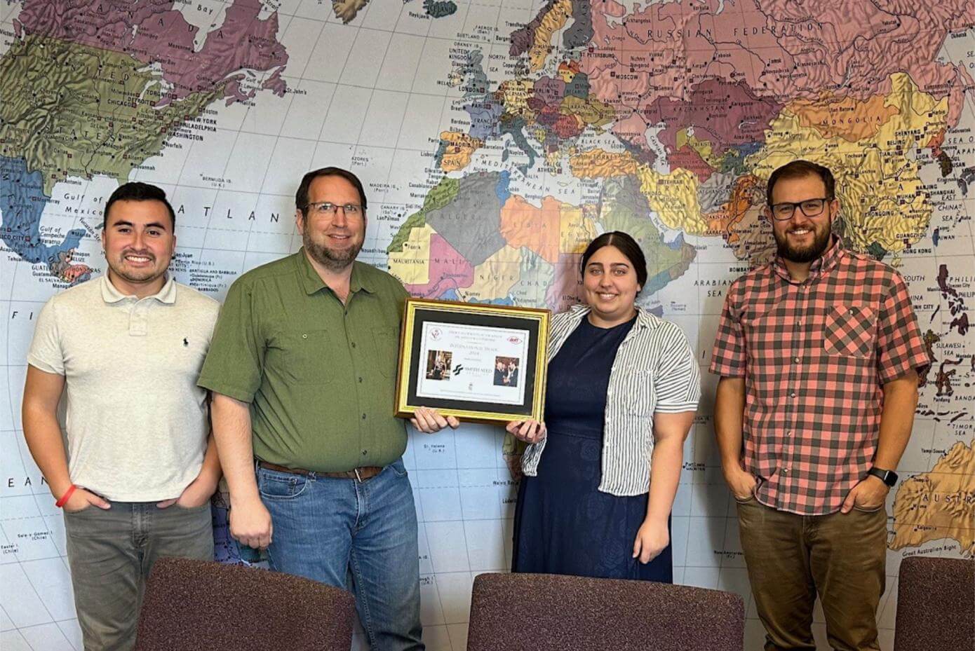 Three men and one woman holding a framed document in front of a world map.