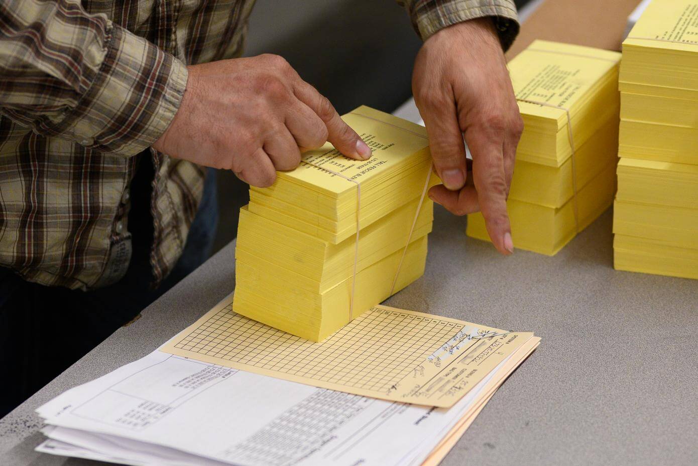 A person wearing a plaid shirt organizes several neat stacks of yellow paper sheets on a table.
